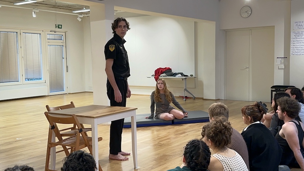 An actor wearing a police uniform stands looking towards the audience while a second actor is seated on mats on the floor in mid-performance of 'Faith Hope Charity'. Audience members watch the performance.